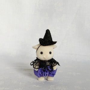 May include: A small, cream-colored flocked animal figurine dressed in a black glittery pointed hat, a black lace collar, and purple fabric bloomers. The figure has a rounded head, small ears, and dark eyes, standing upright in a miniature costume.