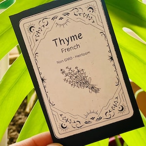 May include: A black and white seed packet with a decorative border. The packet says "Thyme French Non GMO - Heirloom" and features a black and white illustration of a bunch of thyme.
