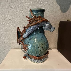 May include: A ceramic vase with a glossy teal glaze and dark speckles, featuring a sculpted three-dimensional dragon wrapped around the neck and body. The dragon has a light blue body with dark blue spots and brown, textured fins and horns.