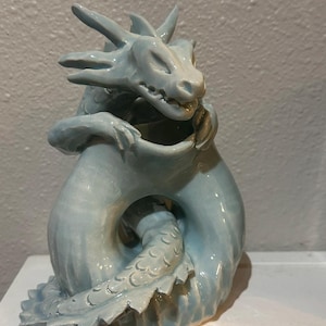 Puede incluir: Una escultura de cerámica de dragón azul claro con una cola enroscada y una cara detallada. El dragón está sujetando su cola con la boca.