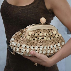 Puede incluir: Un bolso de mano de ganchillo con un patrón de cuadrados de la abuela en tonos beige y crema. El bolso tiene un marco arqueado liso de color claro con un cierre de cuenta de madera redonda en el centro. El cuerpo es de hilo texturizado.