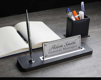 May include: A black leather desk organiser with a silver nameplate that reads "Adam Smith General Manager". A silver pen is in the holder.