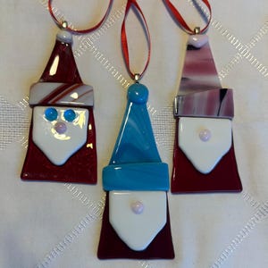 May include: Three glass ornaments shaped like Santa Claus. The ornaments are red, white, blue, and pink. Each ornament has a white beard and a pink nose.