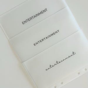 Puede incluir: Tres bolsillos de plástico blanco con la palabra "Entertainment" impresa en cada uno. La palabra está en mayúsculas en los dos bolsillos superiores y en cursiva en el bolsillo inferior.