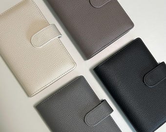 May include: Four leather journals in cream, gray, dark gray, and black. Each journal has a textured surface and a flap closure. The journals are arranged on a white surface.