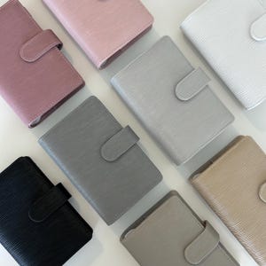 Op de afbeelding: Een collectie van zeven rechthoekige planners met een getextureerd, geribbeld oppervlak, elk voorzien van een bijpassende sluiting met drukknoop. De planners zijn verkrijgbaar in zwart, donkergrijs, lichtgrijs, beige, wit, roze en mauve.