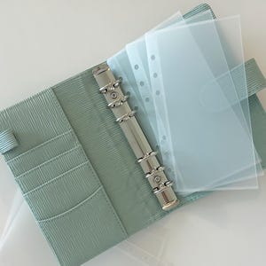 May include: A light green leather bound planner with a silver ring binder and clear plastic page protectors. The planner is open and the page protectors are visible.