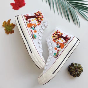 Op de afbeelding: Een paar witte hoge sneakers met gedetailleerd borduurwerk van een oranje vos die een boek leest onder een boom met herfstbladeren, rode paddenstoelen en een stapel kleurrijke boeken. De schoenen hebben witte veters en een dikke rubberen zool.