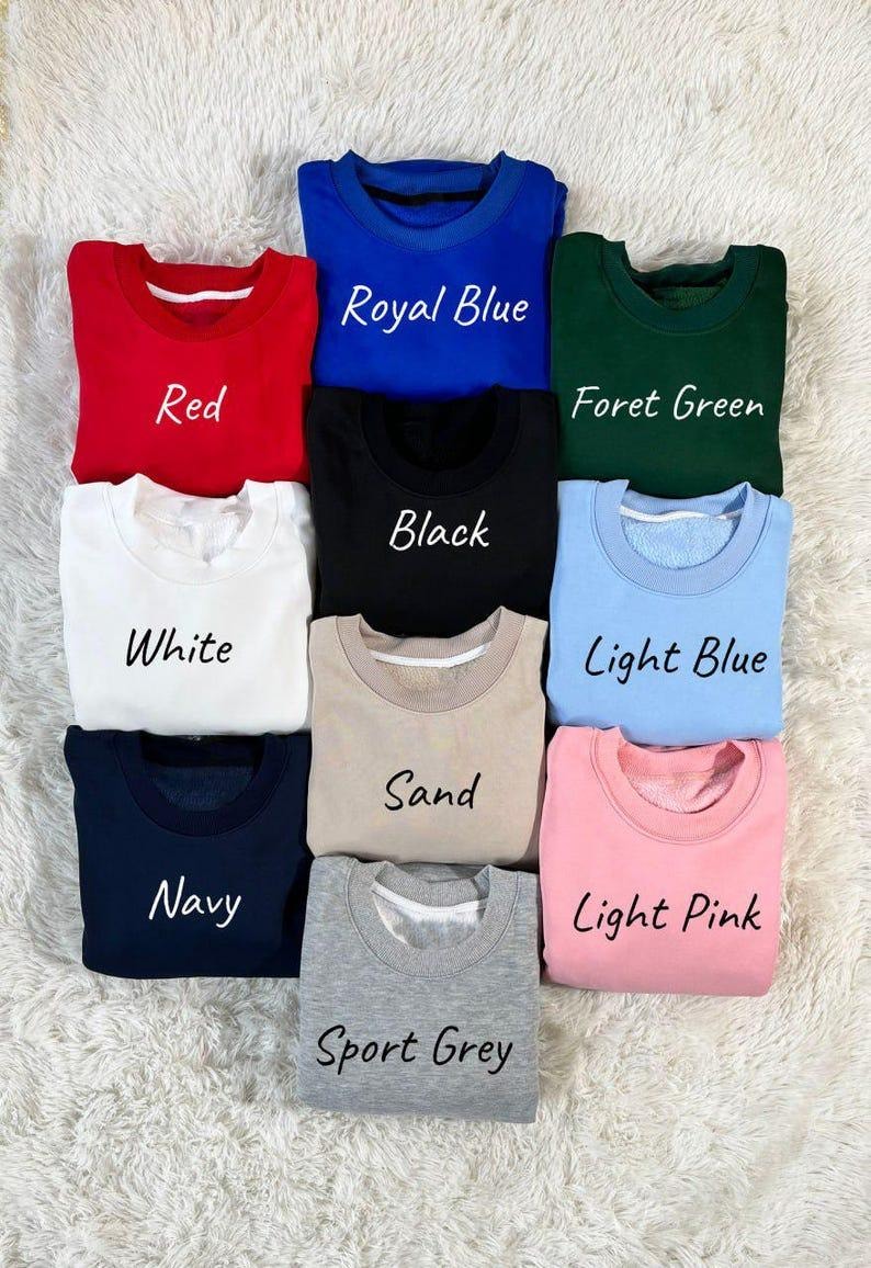 May include: A collection of nine crew neck sweatshirts in various colors, including red, royal blue, forest green, black, white, light blue, sand, navy, light pink, and sport grey. Each sweatshirt has the color name printed on it.