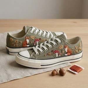 May include: A pair of olive green canvas trainers featuring detailed embroidery of orange foxes, red and white spotted mushrooms, and brown oak leaves. The shoes have white laces, white rubber toe caps, and white soles with black trim.