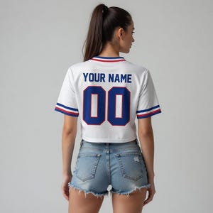 Puede incluir: Camiseta blanca de corte corto con detalles azules y rojos. La parte trasera muestra "YOUR NAME" sobre el n&uacute;mero "00" en azul. Las mangas y el cuello tienen rayas azules y rojas. Se usa con pantalones cortos de mezclilla.