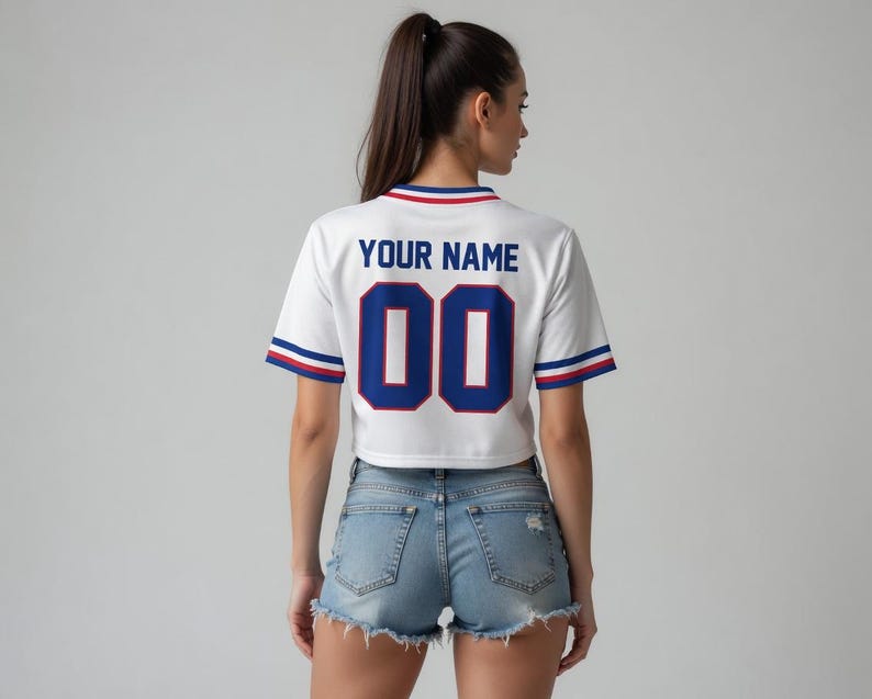 Puede incluir: Camiseta blanca de corte corto con detalles azules y rojos. La parte trasera muestra "YOUR NAME" sobre el n&uacute;mero "00" en azul. Las mangas y el cuello tienen rayas azules y rojas. Se usa con pantalones cortos de mezclilla.