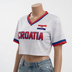 May include: White cropped soccer jersey with red and blue trim. The word "CROATIA" is printed across the front in red block letters. A Croatian flag is on the upper chest. The jersey has a V-neck and short sleeves.