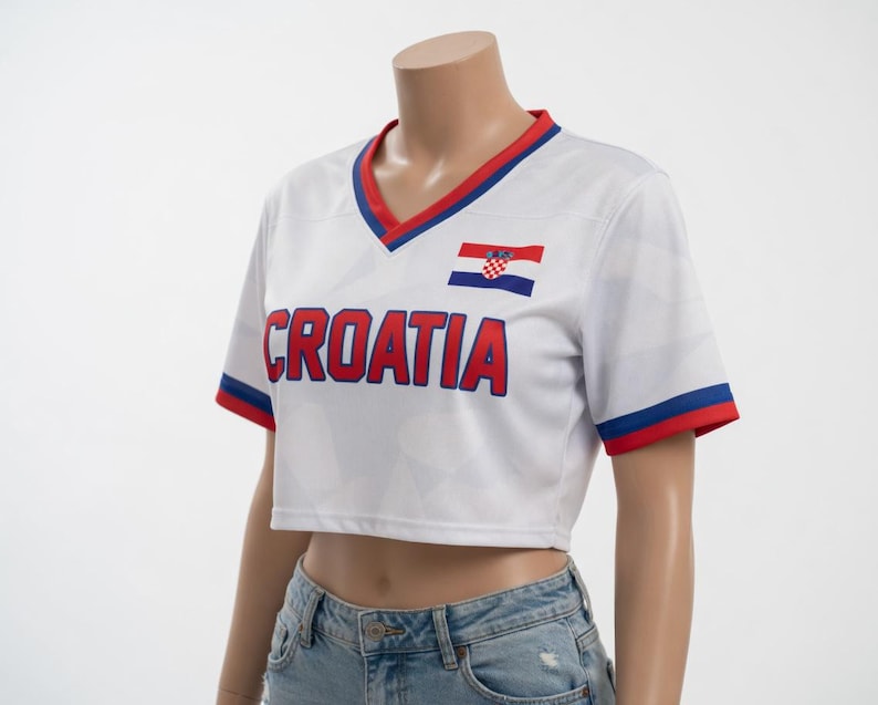 May include: White cropped soccer jersey with red and blue trim. The word "CROATIA" is printed across the front in red block letters. A Croatian flag is on the upper chest. The jersey has a V-neck and short sleeves.
