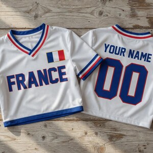 Puede incluir: Dos camisetas deportivas blancas con ribetes azules, rojos y blancos. Una camiseta tiene "FRANCE" impreso en azul, con la bandera francesa. La otra camiseta tiene "YOUR NAME" y el n&uacute;mero "00" en azul.