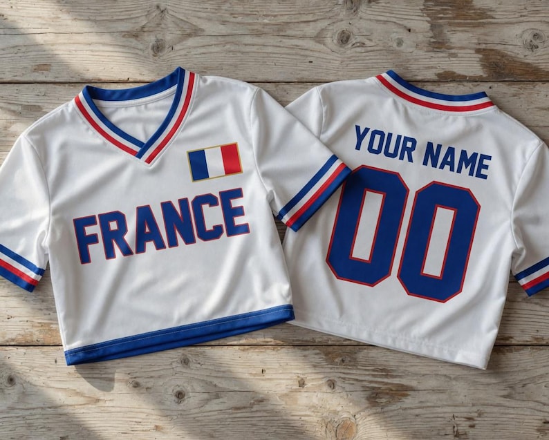 Puede incluir: Dos camisetas deportivas blancas con ribetes azules, rojos y blancos. Una camiseta tiene "FRANCE" impreso en azul, con la bandera francesa. La otra camiseta tiene "YOUR NAME" y el n&uacute;mero "00" en azul.