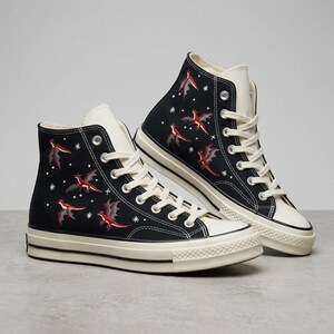Könnte beinhalten: Ein Paar schwarze High-Top-Sneaker aus Segeltuch mit einem gestickten Muster aus kleinen roten Drachen und weißen Sternen. Die Schuhe haben cremefarbene Schnürsenkel, passende Gummikappen und dicke weiße Gummisohlen mit einem dünnen schwarzen Streifen.