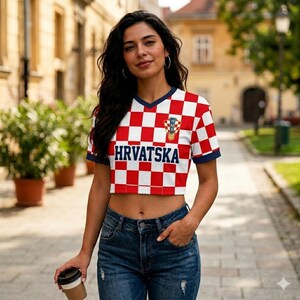 May include: A cropped t-shirt featuring a red and white checkered pattern, a dark blue V-neck collar, and dark blue sleeve cuffs. The word "HRVATSKA" is printed in dark blue across the chest, positioned below a small red, white, and blue emblem.