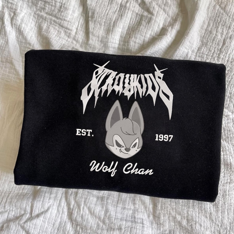 May include: Black sweatshirt featuring a white graphic of the text "Stray Kids" above a cartoon wolf head. Below the wolf head are the words "EST. 1997" and "Wolf Chan" in white. The sweatshirt is folded on a white surface.