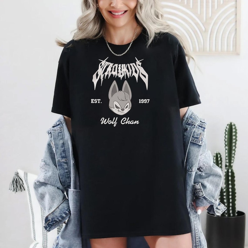 May include: Black t-shirt featuring the text "Stray Kids" in a stylized font above a cartoon wolf face. The text "EST. 1997" and "Wolf Chan" are also printed on the shirt. The shirt is worn with a denim jacket.