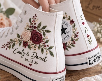 Könnte beinhalten: Ein Paar weiße High-Top-Sneaker aus Segeltuch mit individueller Blumenstickerei in Burgunderrot und zartem Rosa. Die Schuhe sind mit dem Text Emily & Daniel, dem Datum 10.25.26 sowie den Initialen E und D mit einem kleinen roten Herzen personalisiert.