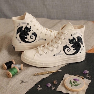 Könnte beinhalten: Ein Paar cremefarbene High-Top-Sneaker aus Segeltuch mit schwarzen gestickten Drachenmotiven, weißen Schnürsenkeln und Gummisohlen. Das Design umfasst kleine gestickte Sterne und Mondsicheln, die die zentralen Drachenfiguren umgeben.