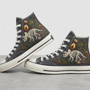 Puede incluir: Un par de zapatillas altas de color gris carbón con cordones blancos y puntera de goma. Los laterales de lona están decorados con esqueletos de triceratops blancos bordados, helechos verdes, follaje marrón y motivos de insectos color ámbar.