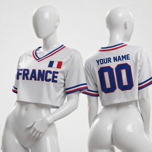 Puede incluir: Camiseta corta blanca con ribetes azules, rojos y blancos. La parte delantera muestra "FRANCE" en letras azules y una bandera francesa. La parte trasera presenta "YOUR NAME" encima del n&uacute;mero "00" en azul.