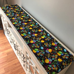 May include: A black table runner with a colorful pattern of balloons and confetti. The text "Happy Birthday" is repeated throughout the pattern.
