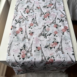 May include: A rectangular table runner featuring a white background with a repeating pattern of black Eiffel Tower illustrations, pink roses, green leaves, butterflies, and circular postmark stamps with the text Paris written in cursive script.