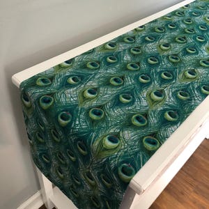 May include: A green and blue peacock feather table runner. The runner is made of fabric and has a repeating pattern of peacock feathers.