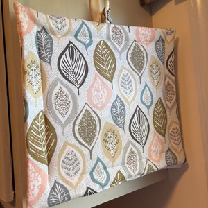May include: A square, fabric kitchen towel with a leaf pattern in shades of pink, green, gray, and white. The towel is hanging from a white hook on a wall. The leaf design is repeated across the surface.