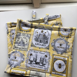 May include: Two yellow and white pot holders with a bee theme. The pot holders feature a honeycomb pattern, bees, and text that says "Honey House", "Honey Farm", "Apiary", "Wild Flower", "Golden Apiaries", and "100% Organic Honey".