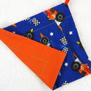 May include: A blue and orange fabric item with a race car theme. The blue side features race cars, checkered flags, and stars. The orange side is solid. The edges are sewn with a green zigzag stitch. A red loop is attached at the top.
