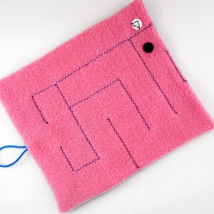 May include: A pink square fabric toy with a maze design stitched in dark blue thread. The toy has a blue loop and is decorated with a black button and a white button with a black triangle.