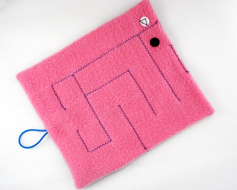 May include: A pink square fabric toy with a maze design stitched in dark blue thread. The toy has a blue loop and is decorated with a black button and a white button with a black triangle.