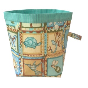 May include: A turquoise fabric storage bin with a patchwork design featuring a turtle, butterfly, hummingbird, and bee. The bin has a handle and a white button closure.
