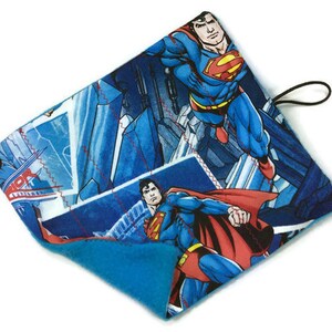 May include: A blue and red Superman-themed item with a black loop. The item features illustrations of Superman in action against a blue cityscape background. The item has a soft, blue interior.