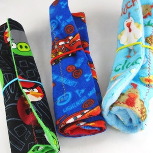 May include: Three rolled fleece blankets, each secured with a rubber band. One features Angry Birds characters on a black background, another has Lightning McQueen from Cars on blue, and the third has chickens on a light blue background.