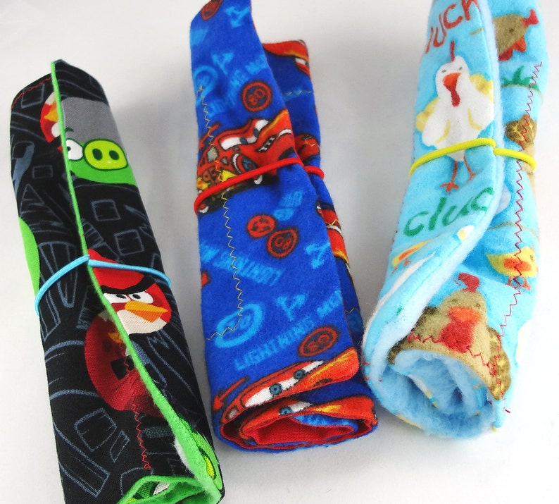 May include: Three rolled fleece blankets, each secured with a rubber band. One features Angry Birds characters on a black background, another has Lightning McQueen from Cars on blue, and the third has chickens on a light blue background.