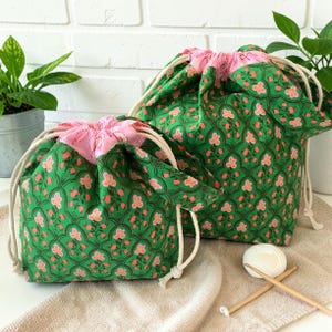 May include: Two fabric drawstring bags featuring a green background with a repeating pattern of pink flowers and dark green scalloped arches. The bags have a light pink interior lining and thick white rope drawstrings for closure.