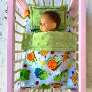 May include: A doll with a smooth head lies in a pink crib with light-colored slats. The doll is covered by a light green textured blanket and rests on a matching pillow. Underneath, a light blue blanket features a pattern of snails and bugs.