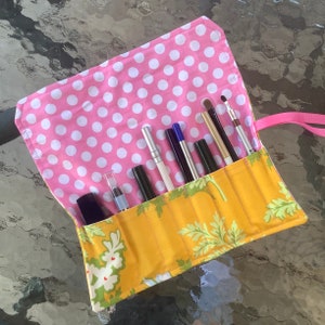 May include: A yellow and pink fabric makeup brush roll with a floral pattern. The roll is open and contains a variety of makeup brushes and pencils.