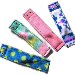 May include: Four fabric wrist straps with different patterns and colours. The straps have two snap buttons on one end. The patterns include blue and green polka dots, pink and green tie dye, pink and blue tie dye, and pink and white tie dye.