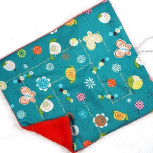 May include: A teal, square-shaped fabric item with a red interior. The teal side features a repeating pattern of butterflies, flowers, and dots in white, orange, yellow, and green. A white loop is attached to the corner.