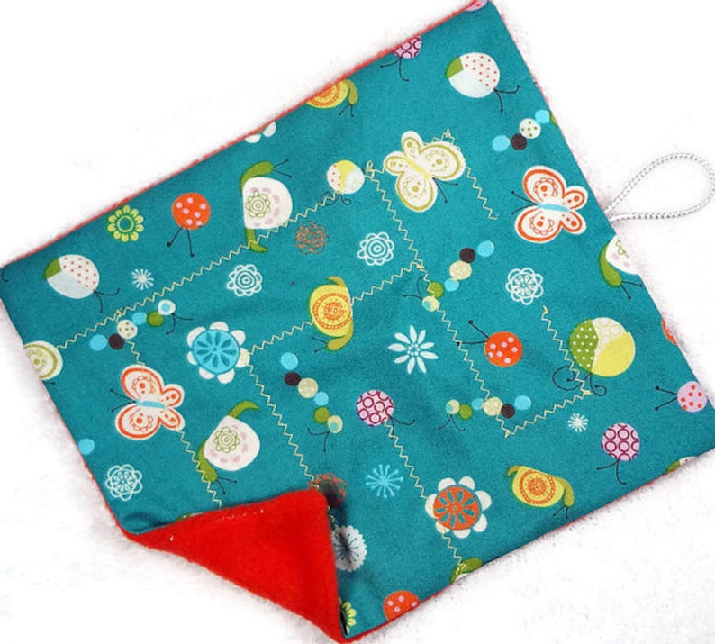 May include: A teal, square-shaped fabric item with a red interior. The teal side features a repeating pattern of butterflies, flowers, and dots in white, orange, yellow, and green. A white loop is attached to the corner.