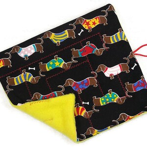 May include: A black pet bed with a repeating pattern of dachshunds wearing colorful sweaters and bone accents. The bed has a bright yellow interior and a red loop for hanging.