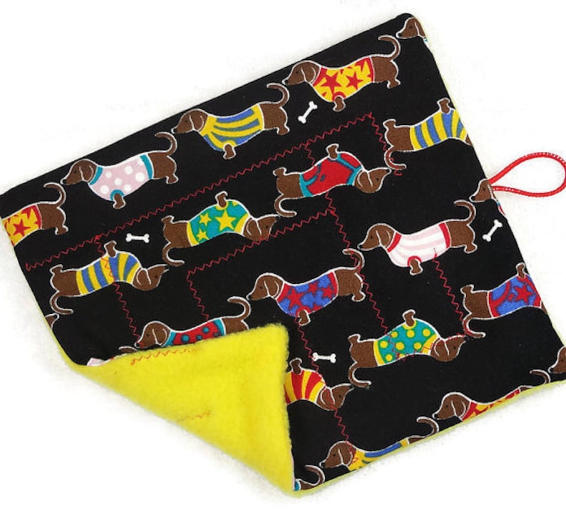 May include: A black pet bed with a repeating pattern of dachshunds wearing colorful sweaters and bone accents. The bed has a bright yellow interior and a red loop for hanging.