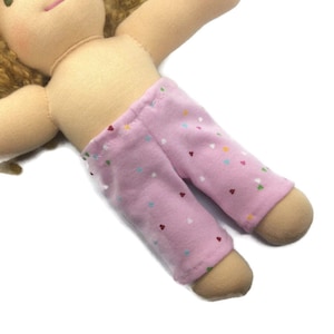 May include: A handmade doll with tan skin and brown hair. The doll is wearing pink trousers patterned with small, colourful hearts. The doll's arms are outstretched, and the legs are slightly bent.