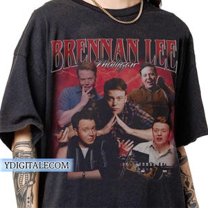 May include: Dark gray t-shirt featuring a graphic of Brennan Lee Mulligan with the text "Brennan Lee Mulligan" in red and white. The design includes multiple images of the person on a red background with lightning bolts.